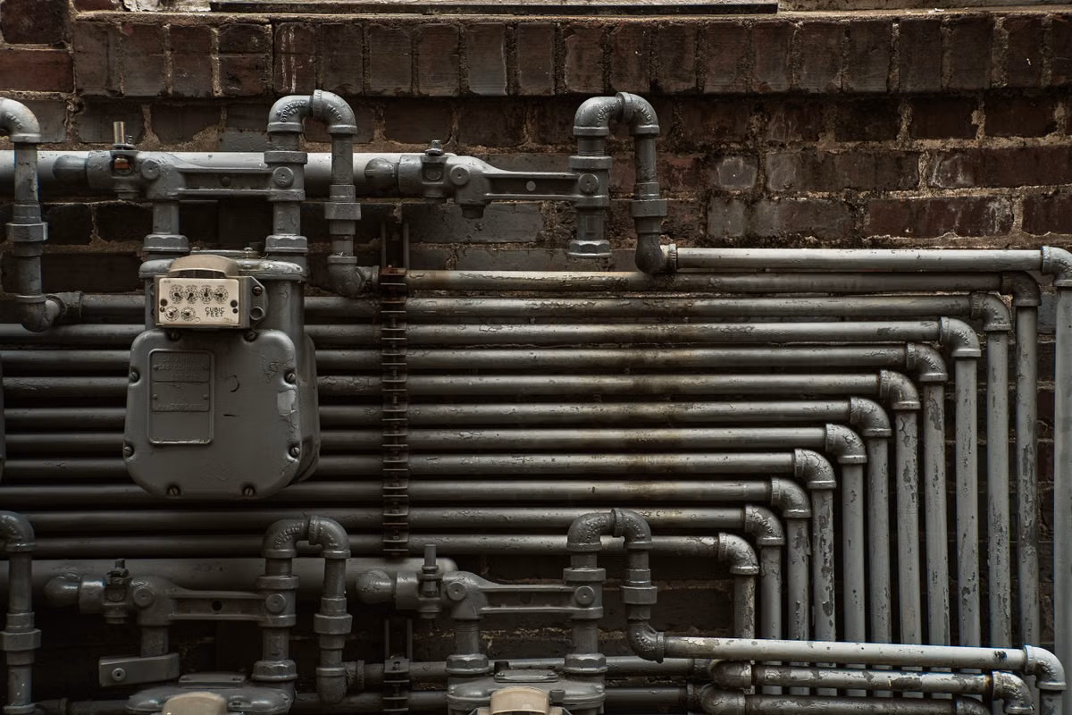 A grey gas meter attached to rows of grey metal pipework running across a dark brick wall.