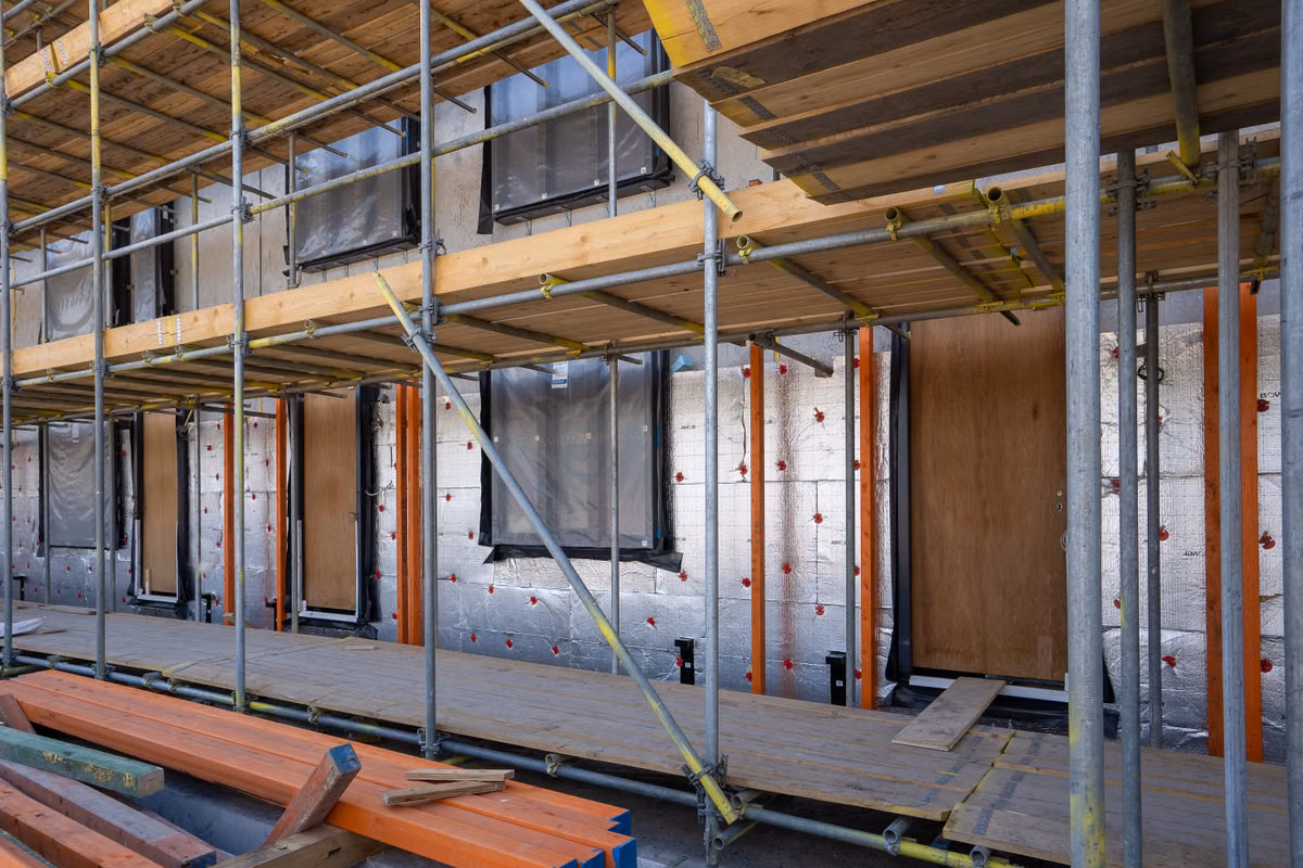 Scaffolding in front of a building wall covered in foil-faced insulation boards, with window openings wrapped in protective sheeting and timber stacked in the foreground.