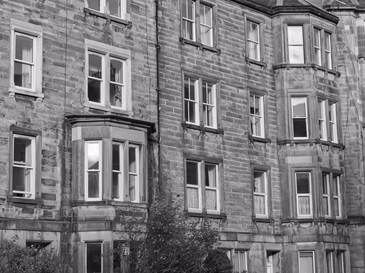 A black and white photograph of a terrace of sandstone tenement flats with bay windows and sash windows.
