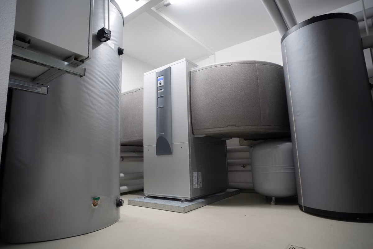 An indoor heat pump unit standing between large insulated hot water and buffer cylinders in a plant room.